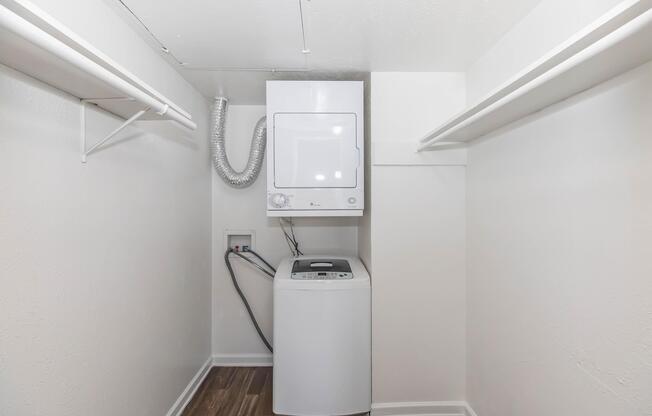 A small laundry room featuring a stacked washer and dryer unit against a plain wall. There are two shelves on either side of the unit, and the floor is covered with laminate flooring. The space is well-lit and uncluttered, with a clean and modern appearance.