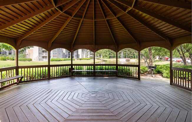 A wooden pavilion with a circular floor and a roof next to a pond and grill