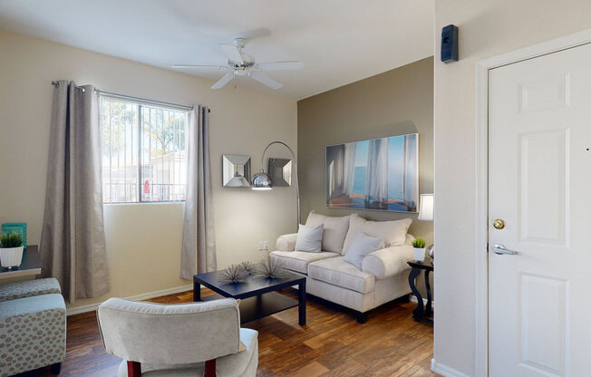 A bright and inviting living room here at Pala Mesa featuring warm wood-style flooring, a plush cream sofa with soft accent pillows, and a sleek dark coffee table centered between cozy seating. A large window with curtains brings in natural light, while modern wall art and a stylish floor lamp create a comfortable and welcoming atmosphere near the entryway.