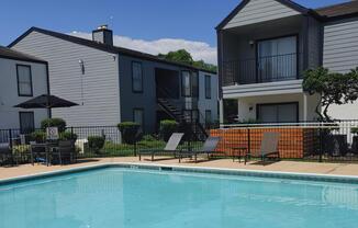 A clear blue swimming pool surrounded by lounge chairs and a shaded umbrella, with two apartment buildings in the background under a bright blue sky. The area is well-maintained with green shrubs and trees.