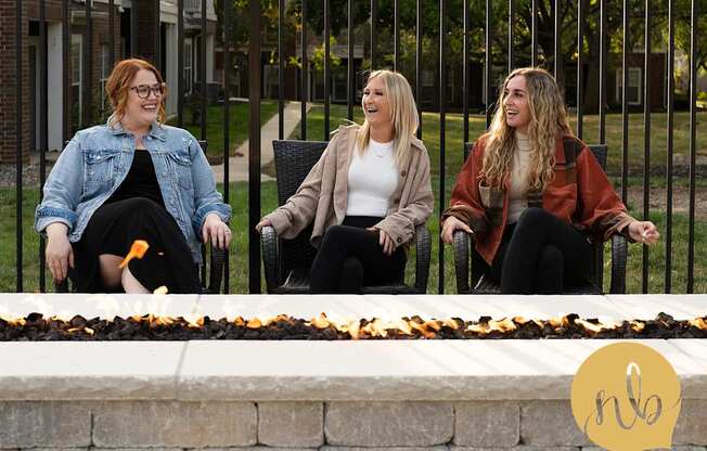 Three women sitting on a bench with a fire pit in front of them.