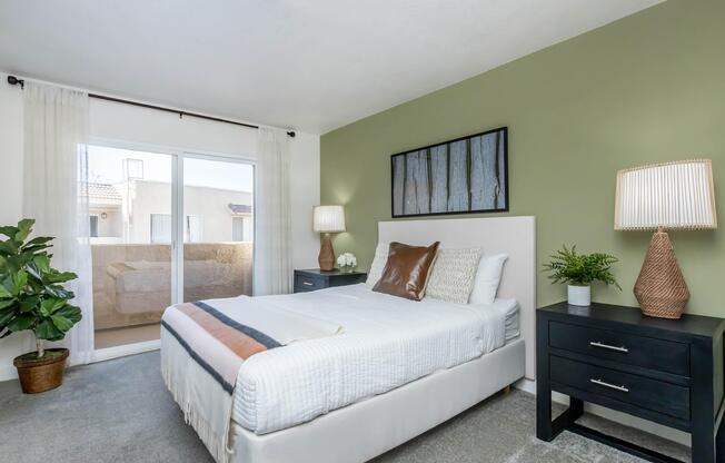 A cozy bedroom featuring a white bed with decorative pillows, two modern bedside tables with lamps, and a potted plant. The walls are painted a soft green, and there is a large window with sheer curtains allowing natural light. A balcony can be seen through the window.