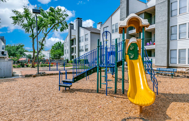 a playground with a yellow slide and blue playset in front of apartments