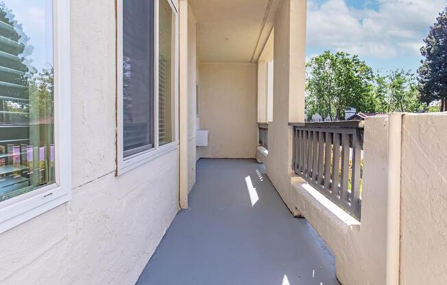 A clean, empty balcony with a light gray floor and white walls. Sunlight filters in through nearby windows, casting soft shadows. The railing is made of wood, and there are green trees in the background, contributing to a serene outdoor atmosphere.