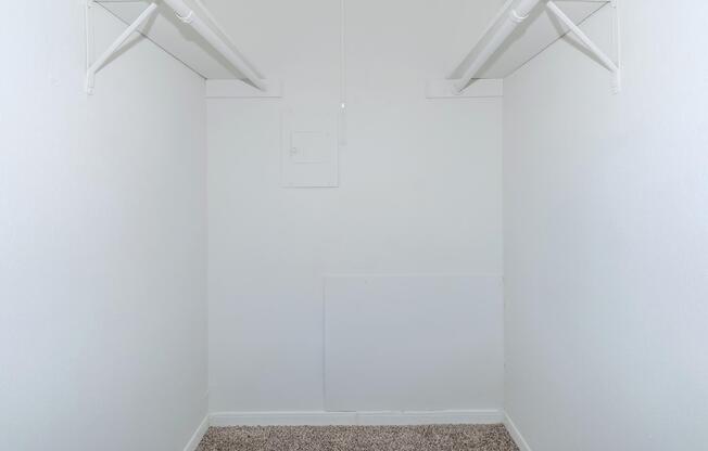 A clean, empty closet with white walls and two overhead clothing rods. The floor is covered with light-colored carpeting, and there is a small electrical panel on the wall to the left. The space is well-lit and appears uncluttered, offering a minimalistic aesthetic.