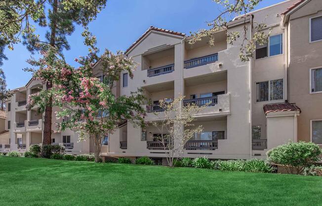 A three-story apartment building with a Mediterranean architectural style, featuring balconies and large windows. The building is surrounded by lush green grass and flowering trees under a clear blue sky, creating a serene and inviting atmosphere.