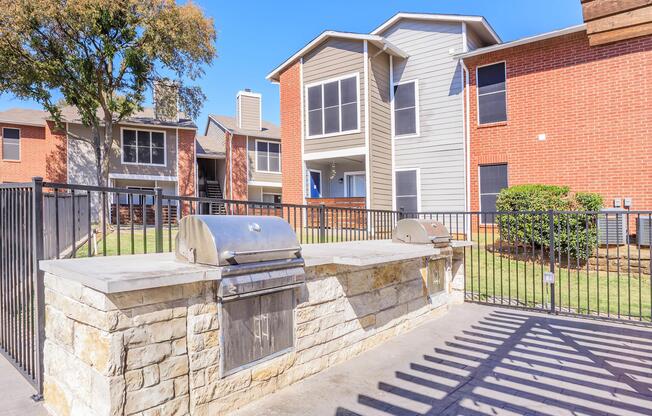 Outdoor grilling area featuring two metal grills on a stone countertop, surrounded by a fenced patio. In the background, there are two residential buildings with a mix of brick and siding, green lawns, and trees, creating a welcoming communal space.