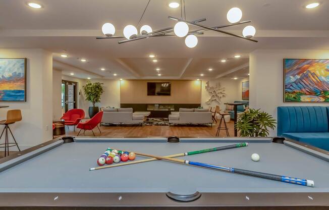 A modern lounge area featuring a pool table with colorful balls and cues arranged on it. In the background, comfortable seating clusters and decorative plants are visible, along with vibrant artwork on the walls and stylish light fixtures overhead. The space is bright and inviting.