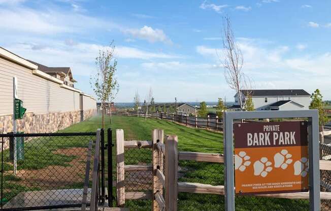 A grassy private bark park here at Prairie Pines at the Ridge featuring a fenced dog-friendly area with a gated entrance, green lawn, young trees, wooden fencing, and a clear bark park sign, offering an open outdoor space for residents to enjoy with their pets.