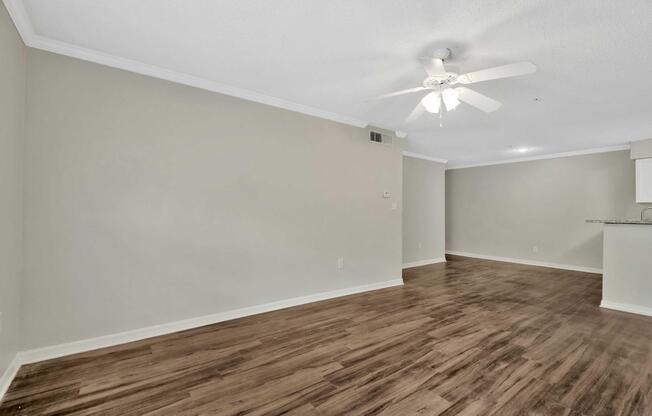 An empty living room with light-colored walls and a ceiling fan. The floor is covered with wooden laminate. There is an open space that leads to a kitchen area in the background, featuring a bar counter. The room is well-lit and appears to be freshly painted with a modern feel.
