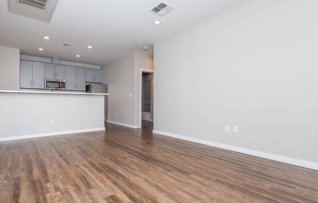 A spacious, modern apartment interior featuring hardwood floors, light grey walls, and an open layout. The kitchen area is visible in the background with light cabinets and stainless steel appliances. There is ample natural light and a door leading to another room.