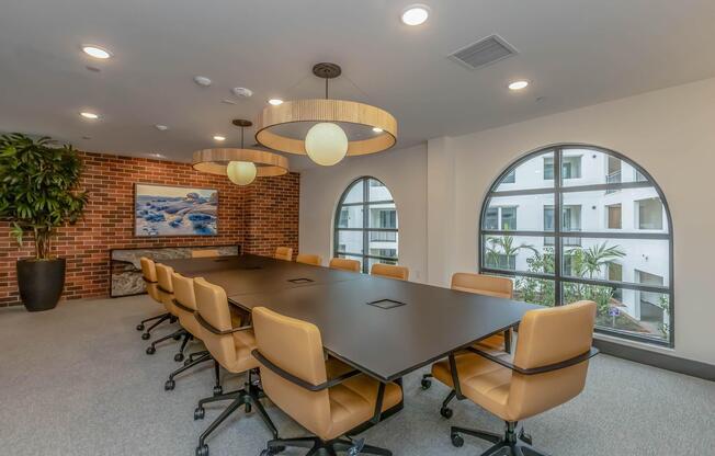 A modern conference room featuring a long black table surrounded by orange swivel chairs. The room has large windows with an outdoor view, a decorative plant, and stylish pendant lighting. A brick wall adds warmth, while a piece of artwork is displayed on the side.