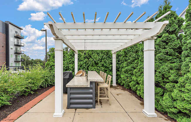 A white pergola with a table and chairs is surrounded by green shrubbery.