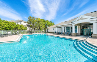 Swimming pool with large tanning deck, table with umbrella, lounge chairs, clubhouse in background surrounded by native landscaping