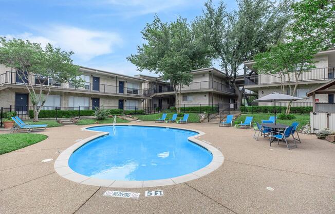 A swimming pool in a courtyard surrounded by residential buildings. The pool features lounge chairs nearby and is marked with a depth sign. Lush greenery is visible around the area, with several trees providing shade. Umbrellas are set up for additional seating comfort.