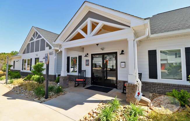 The front of the grey and white clubhouse with green plants and rocking chairs on the porch.