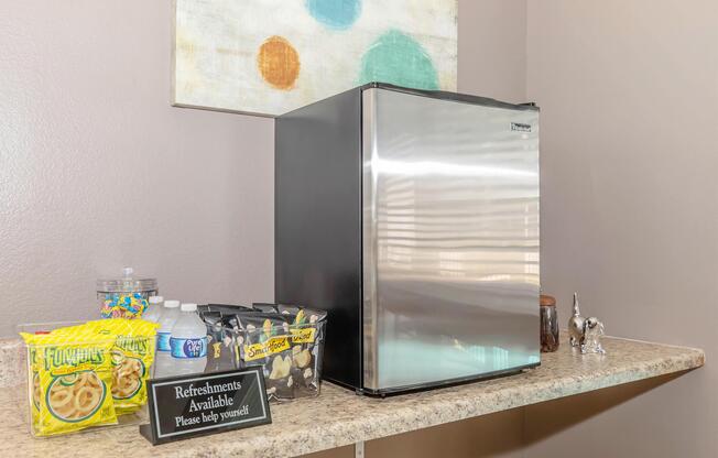 A small stainless steel refrigerator on a countertop, next to a container of snacks, a bottle of water, and a sign that reads "Refreshments Available Please help yourself." The wall behind features an abstract painting with colorful dots.