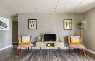 A room with a wooden floor, two chairs, a table with a laptop and a clock.