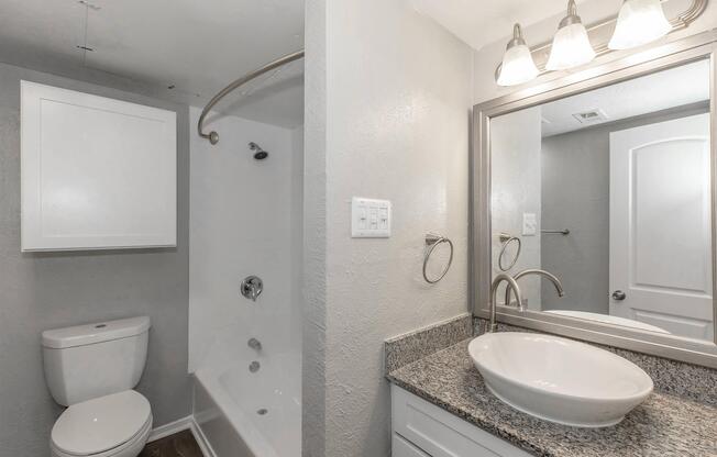 A modern bathroom featuring a white toilet, a bathtub with a shower, and a stylish vanity with a round sink. The walls are painted in a light gray, and there's a mirror above the sink with three light fixtures. Additional storage is provided by a white cabinet.