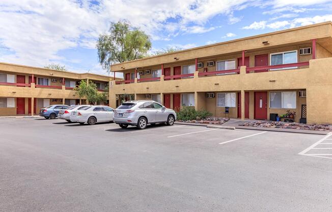 A view of a low-rise motel with two levels, featuring several parked cars in the foreground. The building has a beige exterior with red accents, and there are a few green plants around the entryways. The sky is partly cloudy, indicating a sunny day.