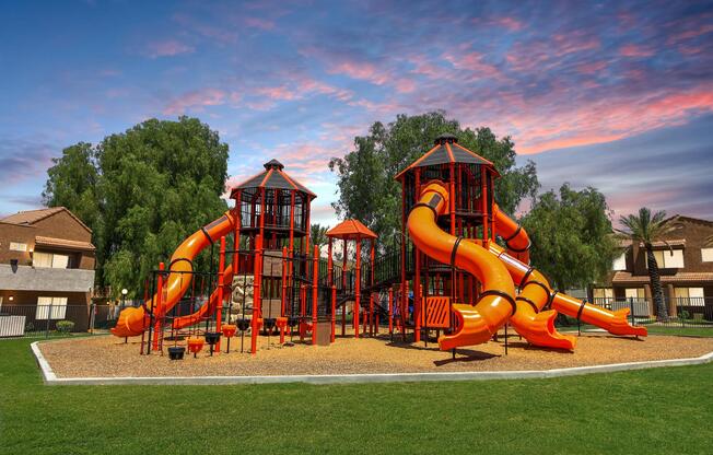 A colorful playground featuring two large play structures with orange slides, surrounded by green grass and trees. The sky is filled with vibrant colors during sunset, creating a cheerful and inviting atmosphere for children to play and explore.