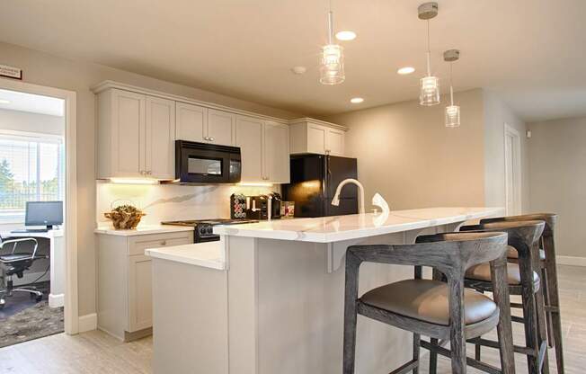 A modern community kitchen here at Andorra showcasing a large island with bar seating, smooth countertops, pendant lighting, and sleek cabinetry, complemented by a built-in microwave, full-size refrigerator, and sink, while recessed ceiling lights and neutral flooring create a bright, inviting space ideal for resident gatherings and shared use.