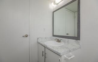 A simple bathroom sink area featuring a white countertop with light gray veining, a round sink, and a bronze faucet. Above the sink is a rectangular mirror framed with a gray border. The bathroom walls are painted white, and there is a door visible in the background.