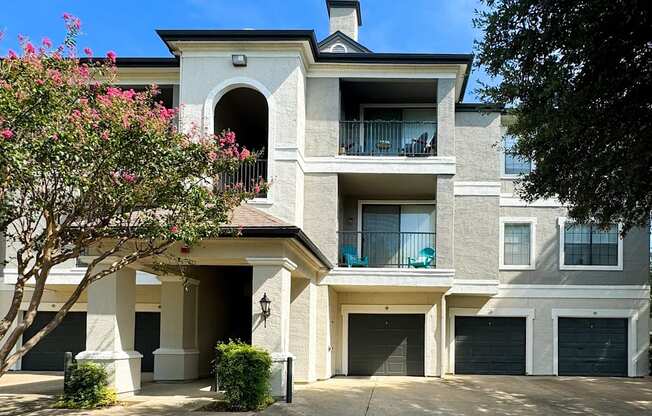 Exterior view of Saxony at Chase Oaks Apartments in Dallas, TX, featuring beige stucco buildings with private balconies, vibrant pink flowering trees, and attached garages.
