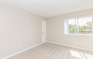 A well-lit, empty room with light beige walls and carpet. There is a white door on one side and a window with sheer curtains allowing natural light. The space appears clean and minimalistic, suitable for various uses such as a bedroom or office.