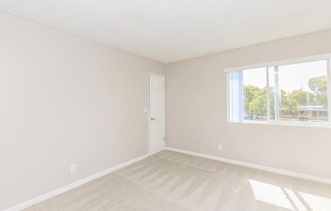 A well-lit, empty room with light beige walls and carpet. There is a white door on one side and a window with sheer curtains allowing natural light. The space appears clean and minimalistic, suitable for various uses such as a bedroom or office.