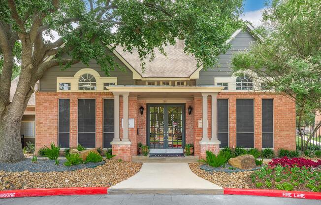 A charming brick and gray house with a covered porch, surrounded by lush greenery and landscaped stones. The entrance features large windows and decorative shutters, with a clear path leading to the door. The property includes well-maintained shrubs and a red curb marking a fire lane.