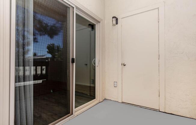 A light-colored, textured wall with a sliding glass door leading to an outdoor area. The door has sheer curtains, and a white door is visible on the right side. The floor is painted gray, creating a simple and clean look. Natural light filters in, giving a bright appearance.