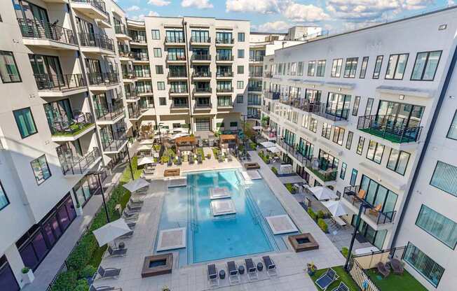 A large swimming pool is surrounded by lounge chairs and umbrellas in a courtyard surrounded by apartment buildings.