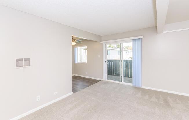 Spacious interior of a light-colored living area with plush carpet, featuring a sliding glass door that opens to a balcony. A ceiling fan is visible, along with a window allowing natural light. The adjacent room is partially visible, enhancing the open layout of the space.
