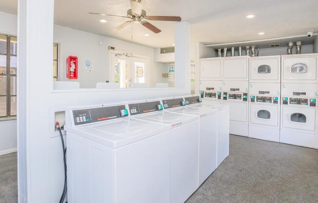 A clean laundry room featuring several white washing machines and dryers, with a ceiling fan above. The room has light-colored walls, windows allowing natural light, and a fire extinguisher mounted on the wall. It creates a well-lit and inviting atmosphere for laundry tasks.
