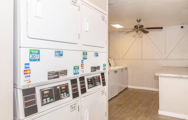 Self-service laundry area featuring several stacked washing machines and dryers with digital control panels. A ceiling fan provides airflow in the room, which has a clean and modern design with light-colored walls and wood-style flooring.
