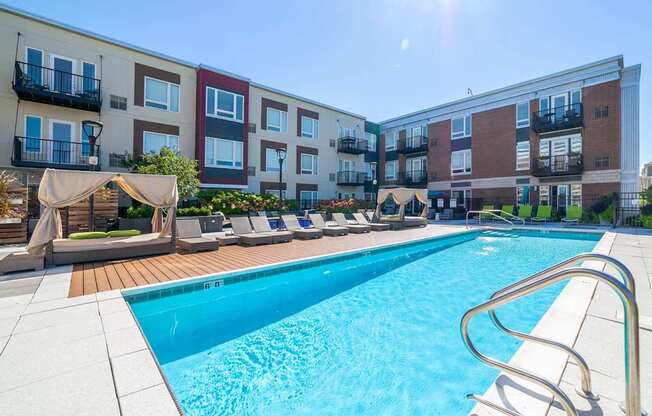 A swimming pool surrounded by lounge chairs and umbrellas in front of apartment buildings.