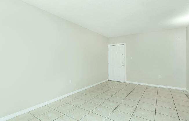 Empty room with light gray walls and a white door. The floor is covered with beige tiles, and there is ample natural light. The space is uncluttered, creating an open and minimalist atmosphere.