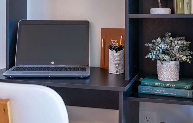 A laptop is on a desk with a white chair, a pencil holder, and a book titled "Brightwood" on a shelf at the Mastlight.