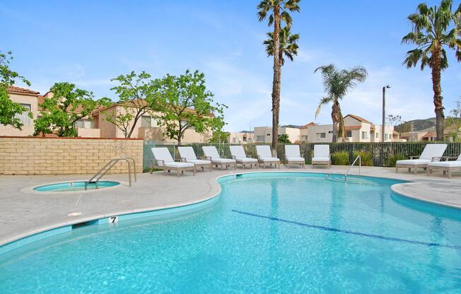 A clear blue swimming pool with a curved edge, surrounded by lounge chairs. Palm trees and green foliage are visible in the background, alongside residential buildings. The sky is bright and sunny, creating a relaxing atmosphere.