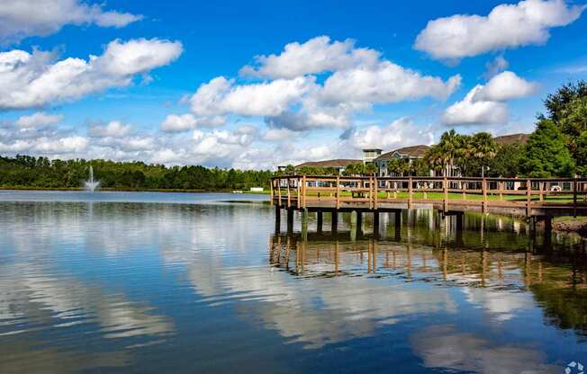 A wooden pier extends into a calm body of water with a fountain in the distance.