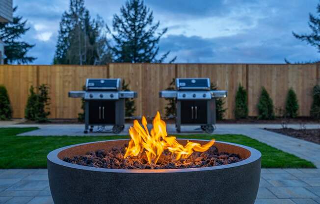 a fire pit in a backyard with two bbq grills in the background
