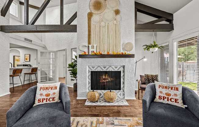 A living room with a fireplace and two grey armchairs with pillows that say pumpkin spice.