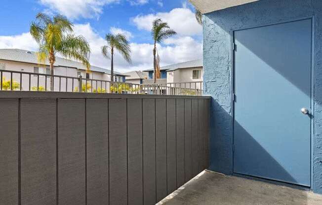 A blue door is on the right side of a wall with a balcony in the background.