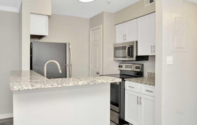 Modern kitchen featuring granite countertops, stainless steel appliances including a refrigerator, stove, and microwave. The cabinetry is sleek and white, while the wall is painted a soft neutral color, creating a bright and inviting atmosphere. A bar area is also visible, connecting to the living space.