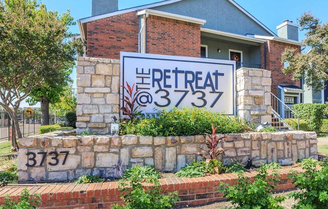 Sign for "The Retreat @ 3737," featuring bold lettering on a large white background, set against a stone and red brick facade. The entrance is landscaped with green plants and colorful flowers, and there are steps leading up to the building in a sunny outdoor setting.