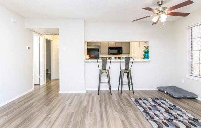 A bright, modern living space featuring a light gray wall, wooden flooring, and a small area rug. Two bar stools are positioned at a kitchen counter with a visible microwave and a blue decorative vase. A fan hangs from the ceiling, and a hallway leads to another room, creating an open and inviting atmosphere.