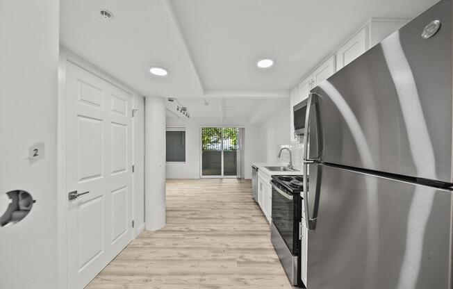 Modern kitchen featuring stainless steel appliances, including a refrigerator and gas stove. The space has white cabinetry and light wood flooring. There are two doors visible, and large windows allow natural light to fill the room, creating a bright and airy atmosphere.