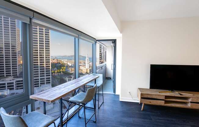 A modern living room with a long wooden table and chairs, a flat screen TV, and a view of the city through the windows.