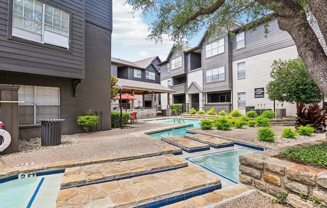 A swimming pool surrounded by stone and a tree in front of apartment buildings.